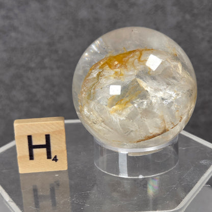 A clear quartz crystal sphere with golden-brown inclusions, displayed on a clear acrylic stand next to a wooden Scrabble tile showing the letter H4, set against a dark gray textured background.