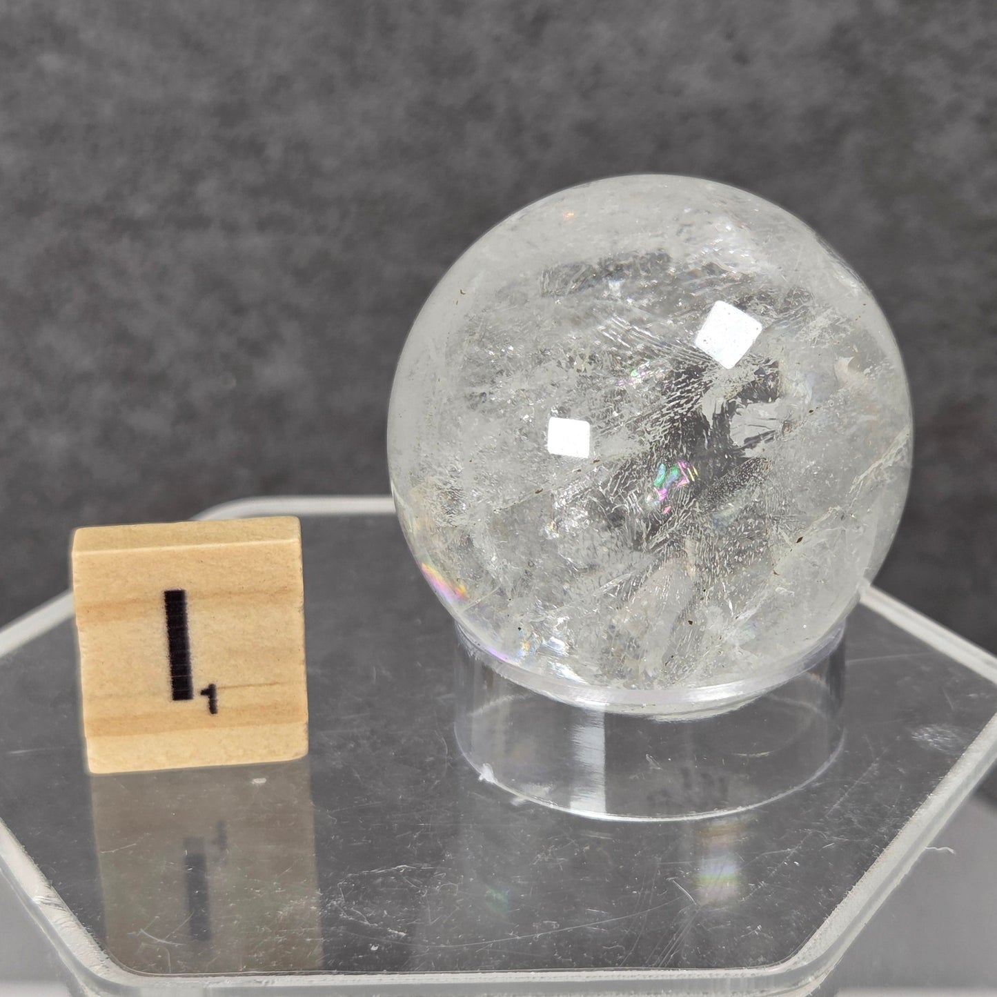 A clear quartz crystal sphere with internal inclusions, displayed on a clear acrylic hexagonal stand next to a small wooden block showing the number one, against a dark gray textured background.