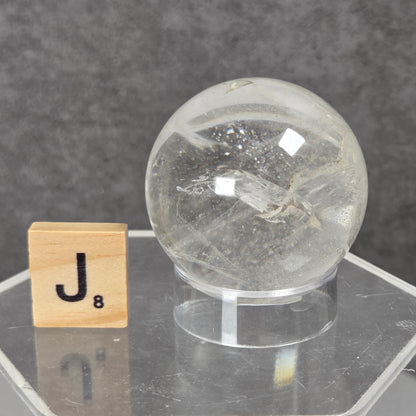 A clear glass or acrylic decorative sphere with internal crystalline texture, displayed on a transparent acrylic stand next to a wooden Scrabble tile showing the letter J, against a dark gray textured background.