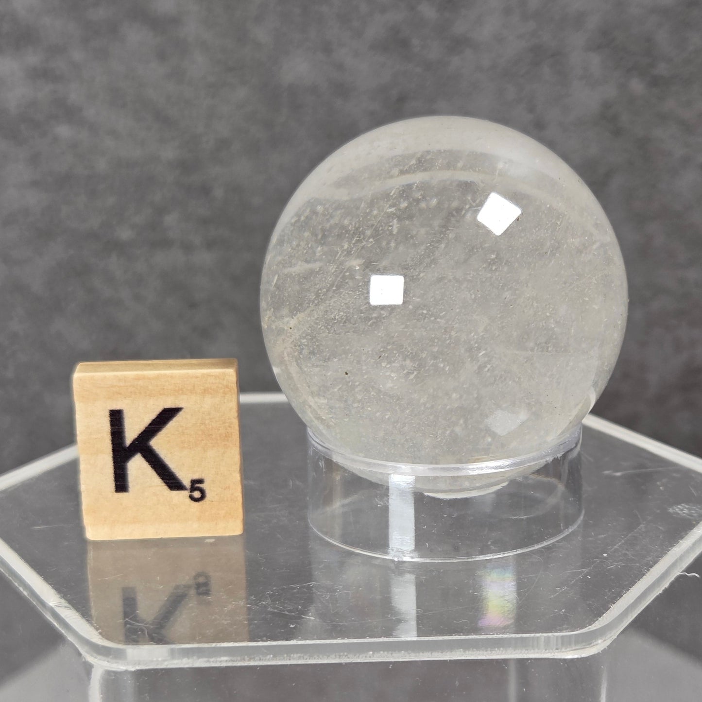 A clear quartz crystal sphere on a clear acrylic display stand, shown next to a wooden Scrabble tile with the letter K on a dark gray textured background.
