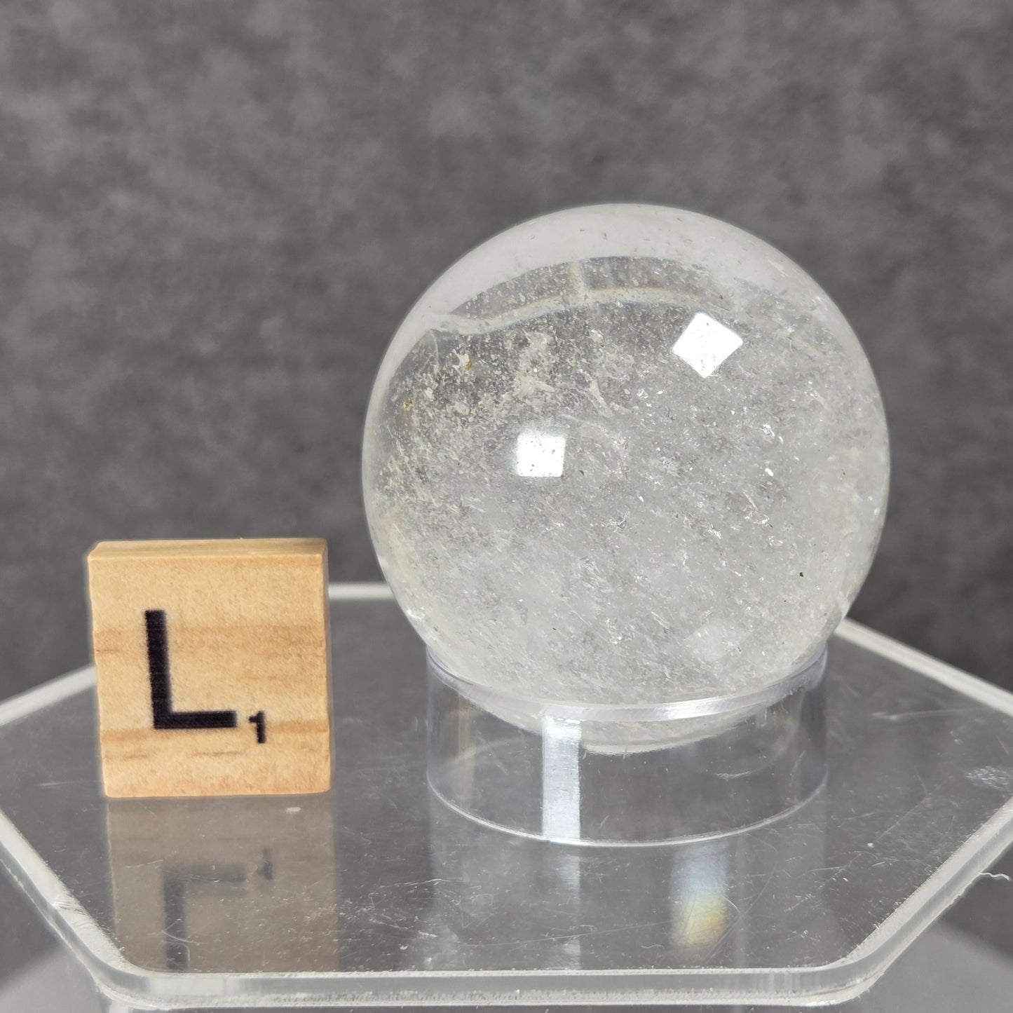 A clear quartz crystal sphere on a clear acrylic stand, shown next to a wooden Scrabble tile with the letter 'L' on a dark gray textured background.