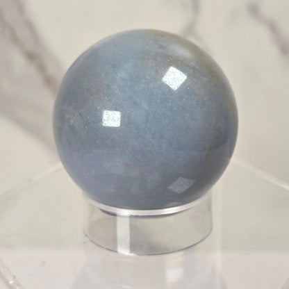 A polished blue-gray stone sphere displayed on a clear acrylic stand against a light marble-patterned background.