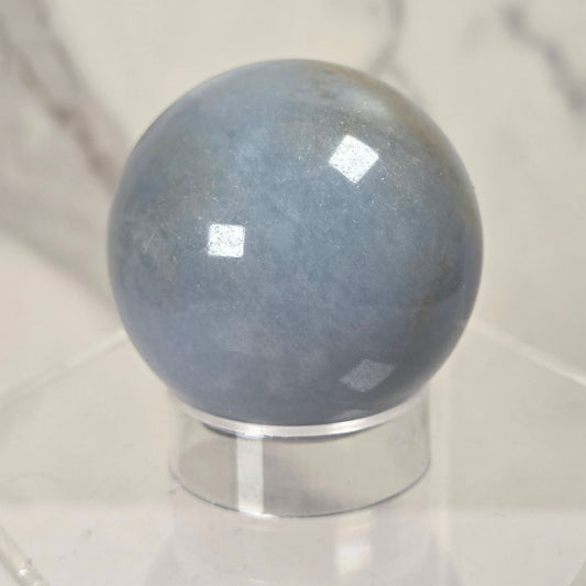 A polished blue-gray stone sphere displayed on a clear acrylic stand against a light marble-patterned background.