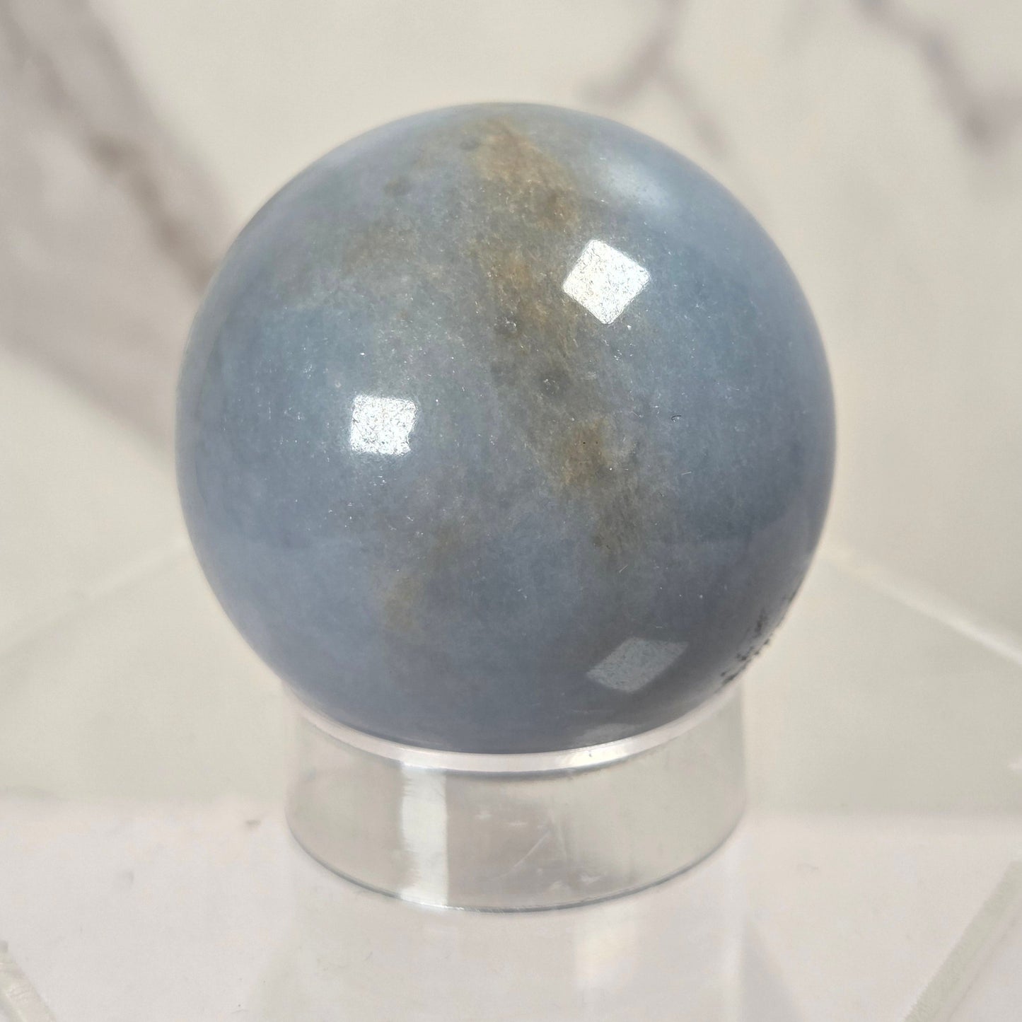 A polished blue-gray stone sphere with subtle brownish veining, displayed on a clear acrylic stand against a light marble-patterned background.