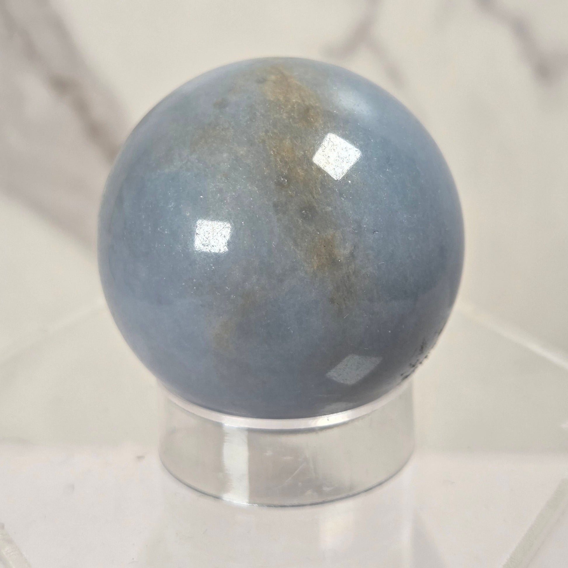 A polished blue-gray stone sphere with subtle brownish veining, displayed on a clear acrylic stand against a light marble-patterned background.