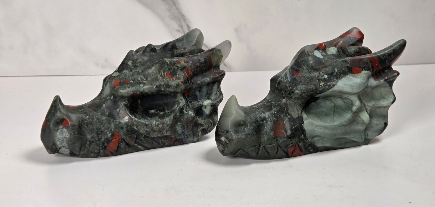 A pair of carved dragon skulls made from polished jasper stone, featuring a mix of dark gray, black, green, and red patterns, displayed on a white surface against a white marble-patterned background.
