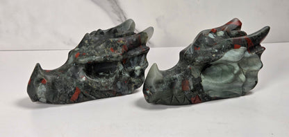 A pair of carved dragon skulls made from polished jasper stone, featuring a mix of dark gray, black, green, and red patterns, displayed on a white surface against a white marble-patterned background.