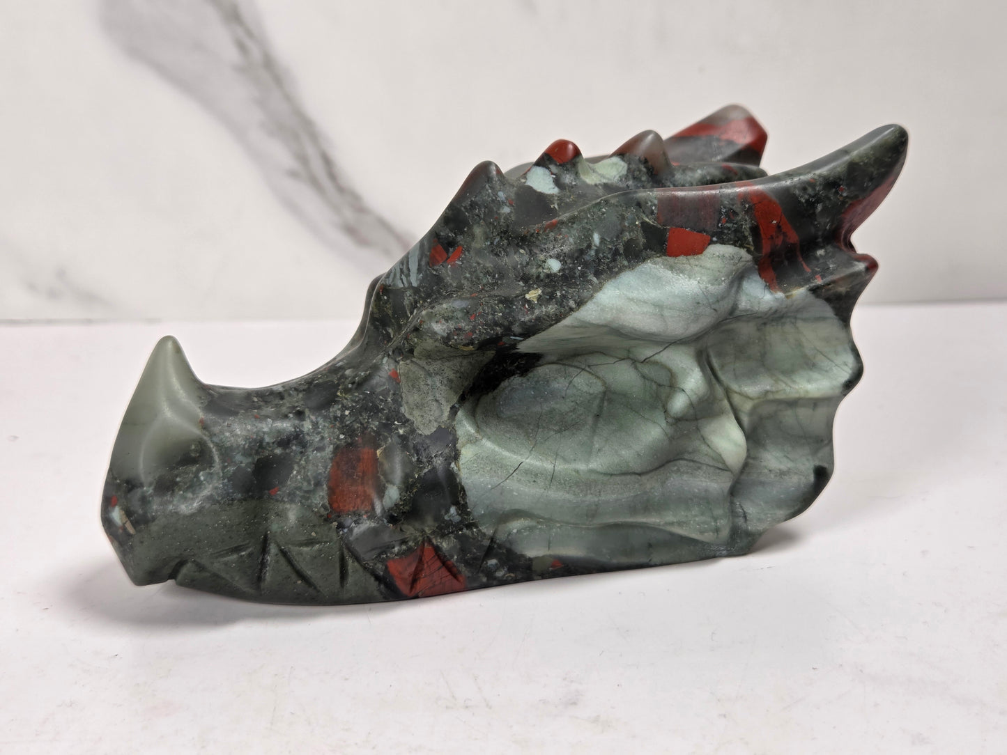 A carved stone dragon head sculpture with a polished finish, featuring shades of gray, black, and red, displayed on a light marble surface.