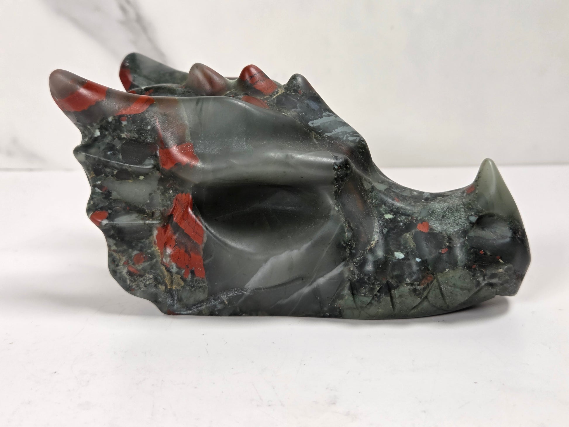A carved stone dragon skull sculpture with a dark gray and black base, accented by vibrant red inclusions and a pale greenish-gray tip, displayed on a light surface.