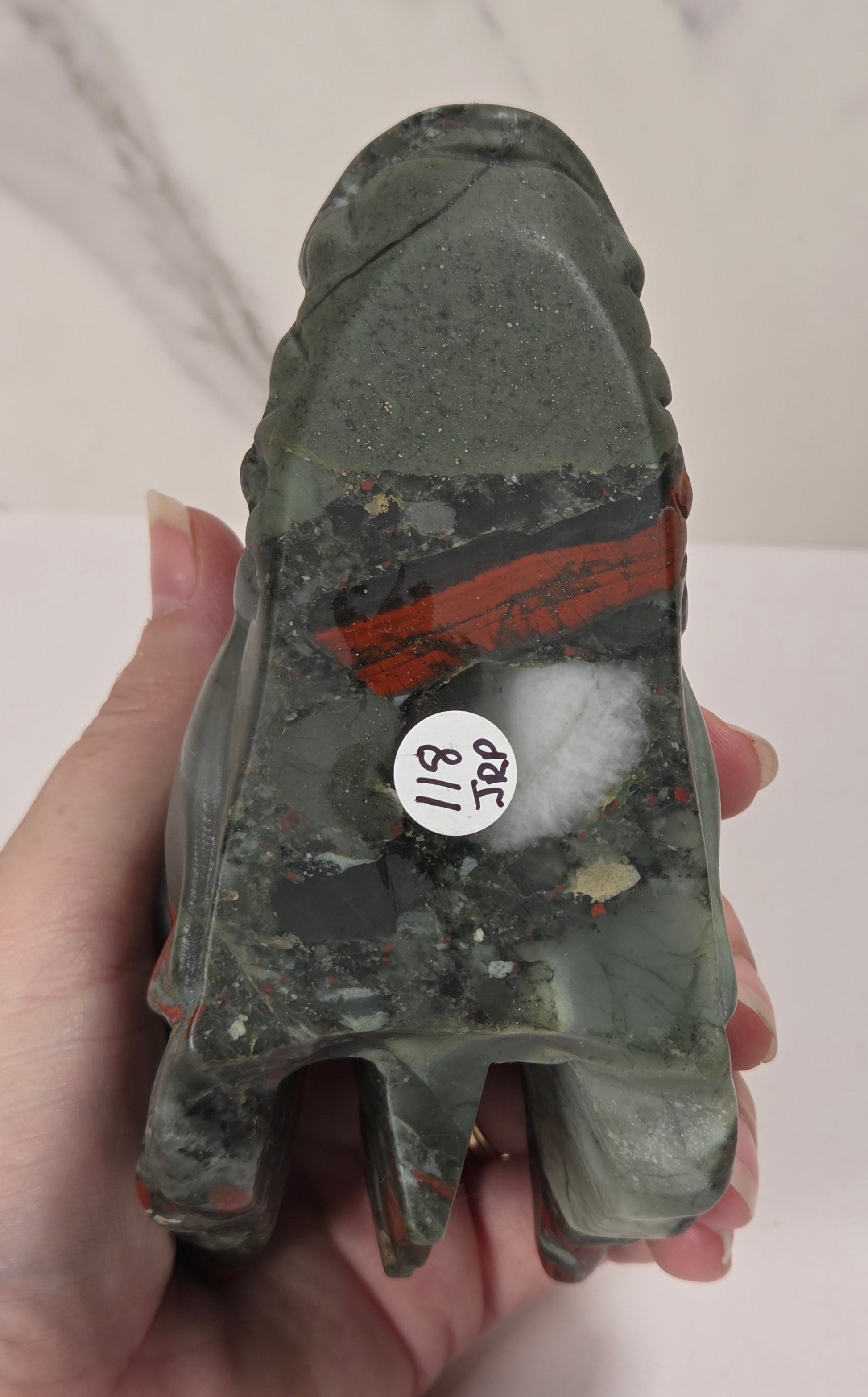 A hand holds a carved stone object, likely a decorative figurine or tool, showing its rough natural texture and color. The stone is primarily dark gray with a prominent reddish-brown stripe and a small white sticker with black text on the side.