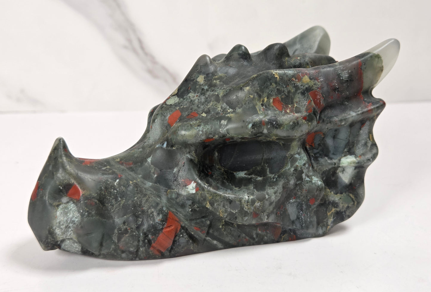 A carved stone dragon head sculpture with a textured, polished finish, featuring a dark gray and black base with vibrant red and orange inclusions, set against a light gray marble-patterned background.