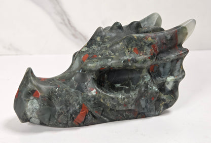 A carved stone dragon head sculpture with a textured, polished finish, featuring a dark gray and black base with vibrant red and orange inclusions, set against a light gray marble-patterned background.