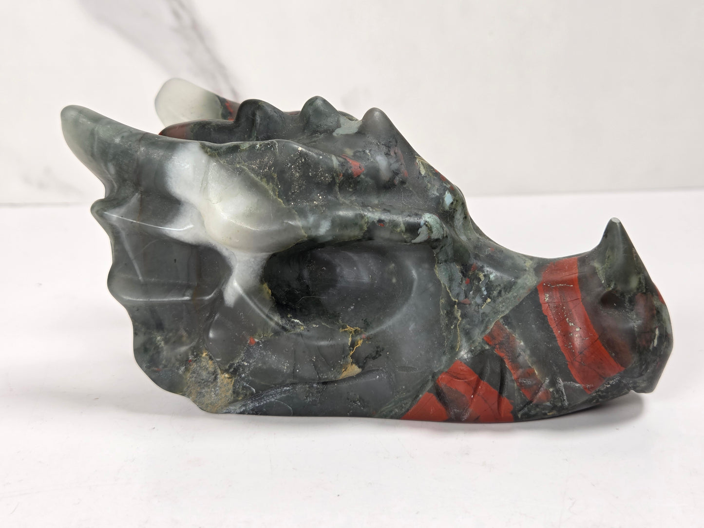 A carved dragon skull figurine made from polished dark gray stone with red and white inclusions, displayed on a light gray surface.
