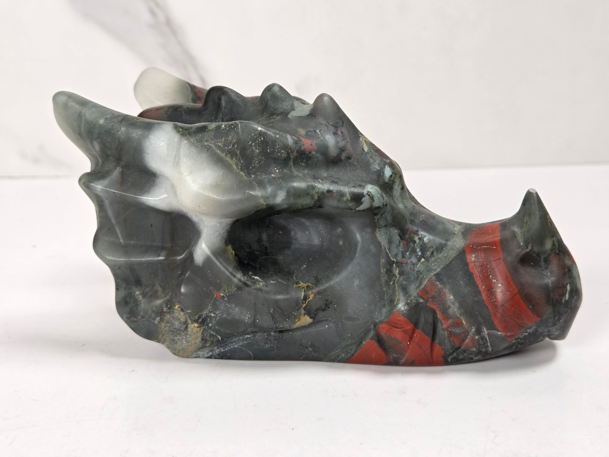 A carved dragon skull figurine made from polished dark gray stone with red and white inclusions, displayed on a light gray surface.