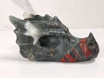 A carved dragon skull figurine made from polished dark gray stone with red and white inclusions, displayed on a light gray surface.