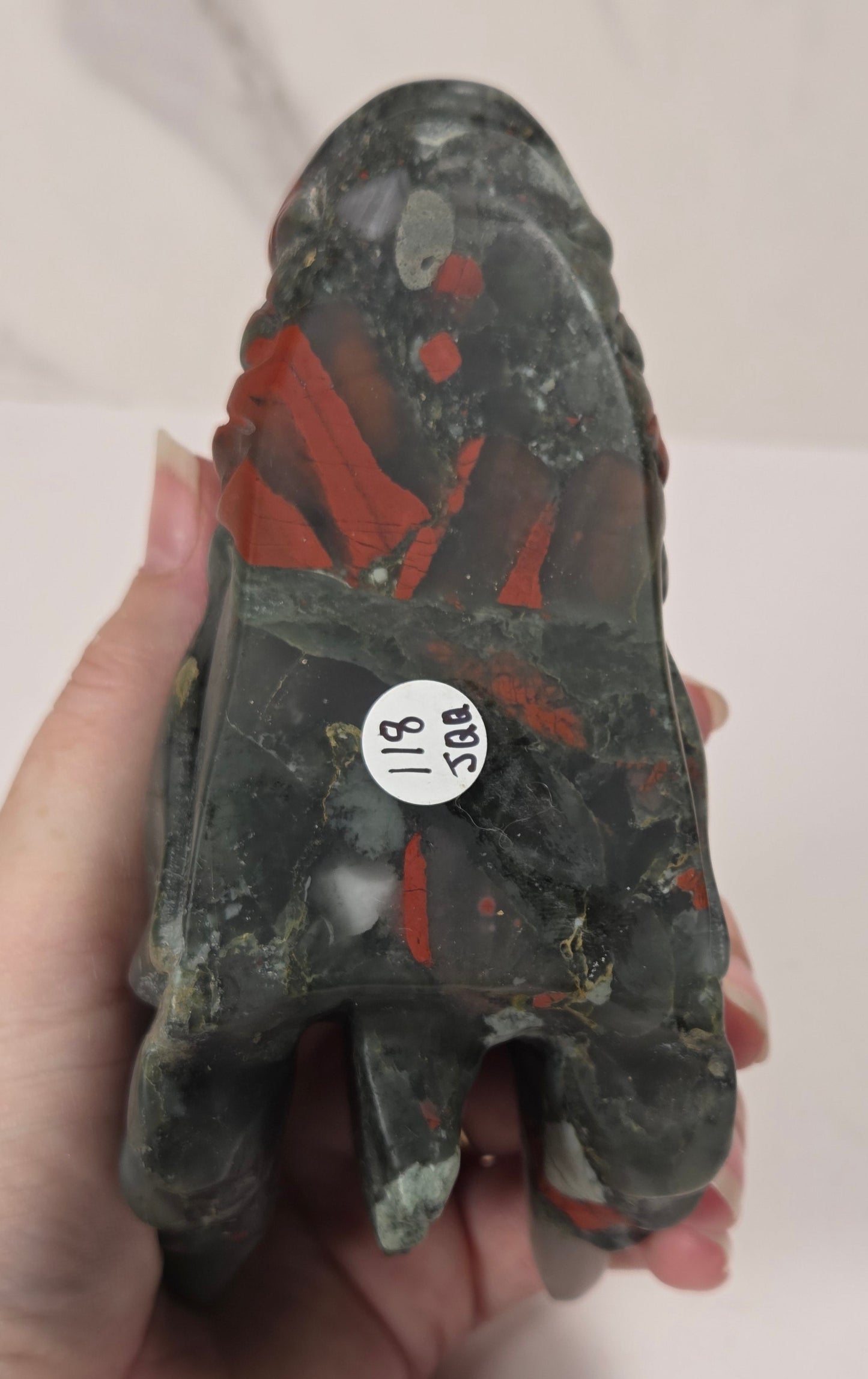 A hand holding a small carved figurine or stone object, showing its dark textured surface with red painted areas and a white sticker with black numbers on the side. The background is a light off-white surface.