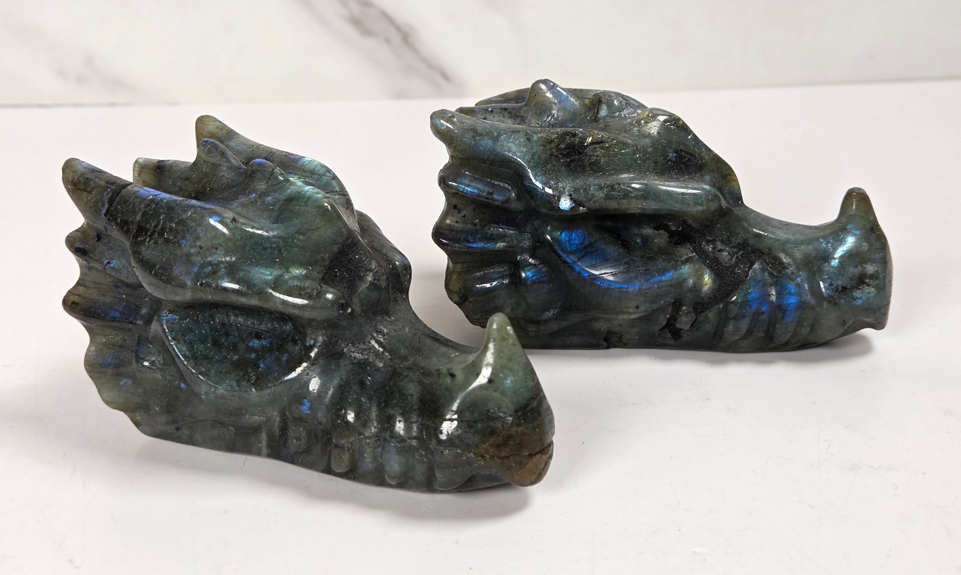 Two small dragon figurines carved from iridescent labradorite, showing blue, green, and gray tones, displayed on a white marble surface.