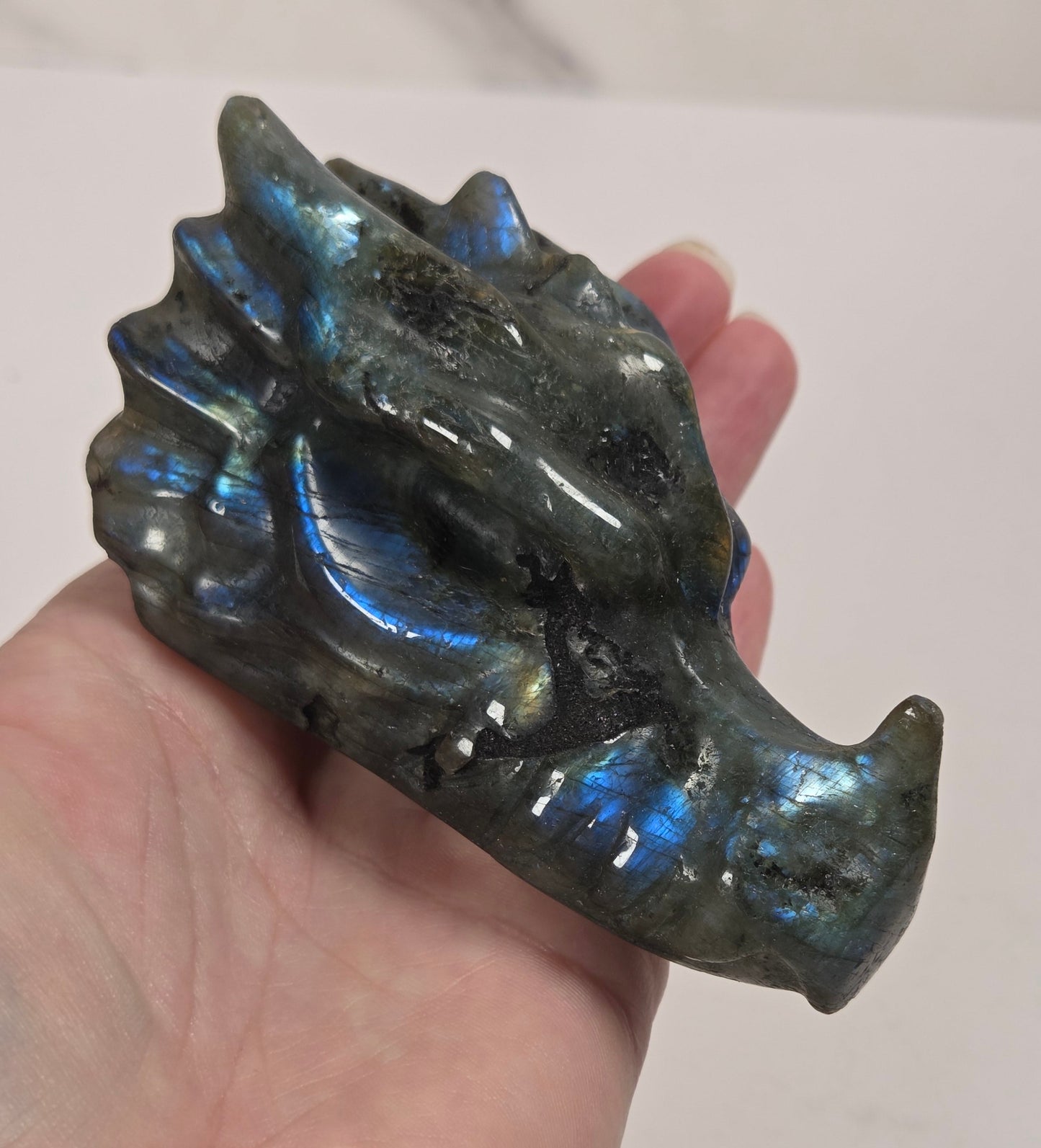 A hand holding a polished labradorite dragon head figurine, showcasing its characteristic blue and green iridescent flashes against a plain light background.