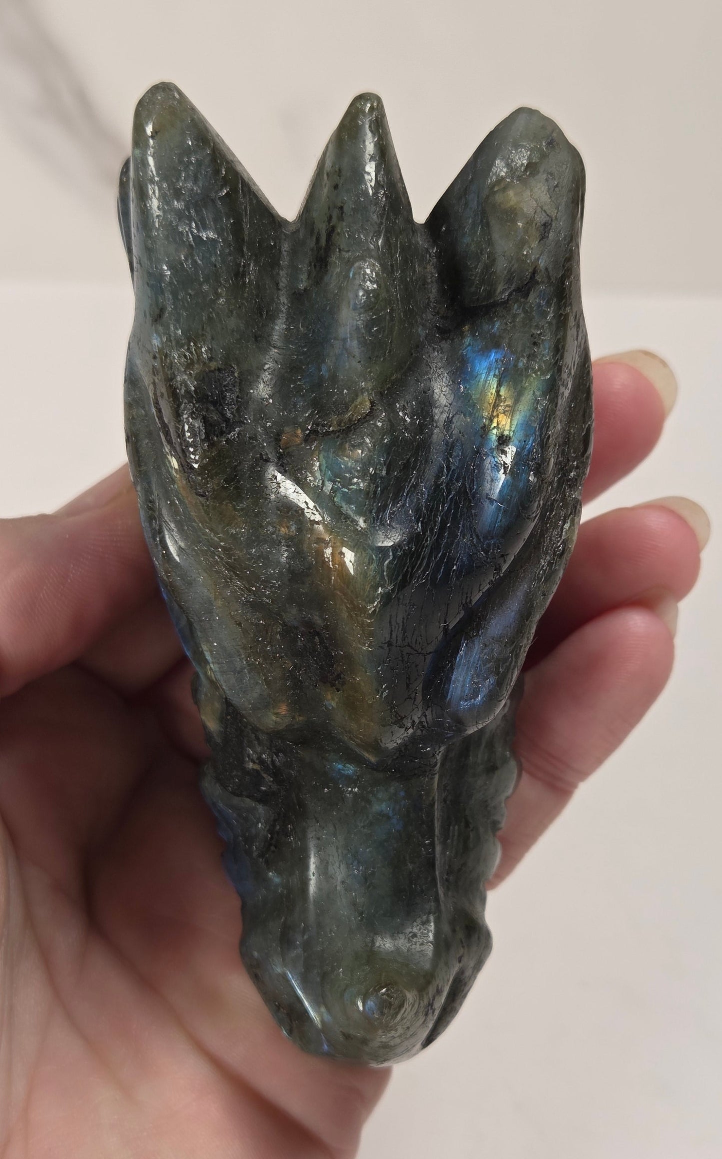 A hand holding a polished Labradorite crystal carving shaped like a horse head, showing its characteristic blue and green iridescent flashes on a light background.
