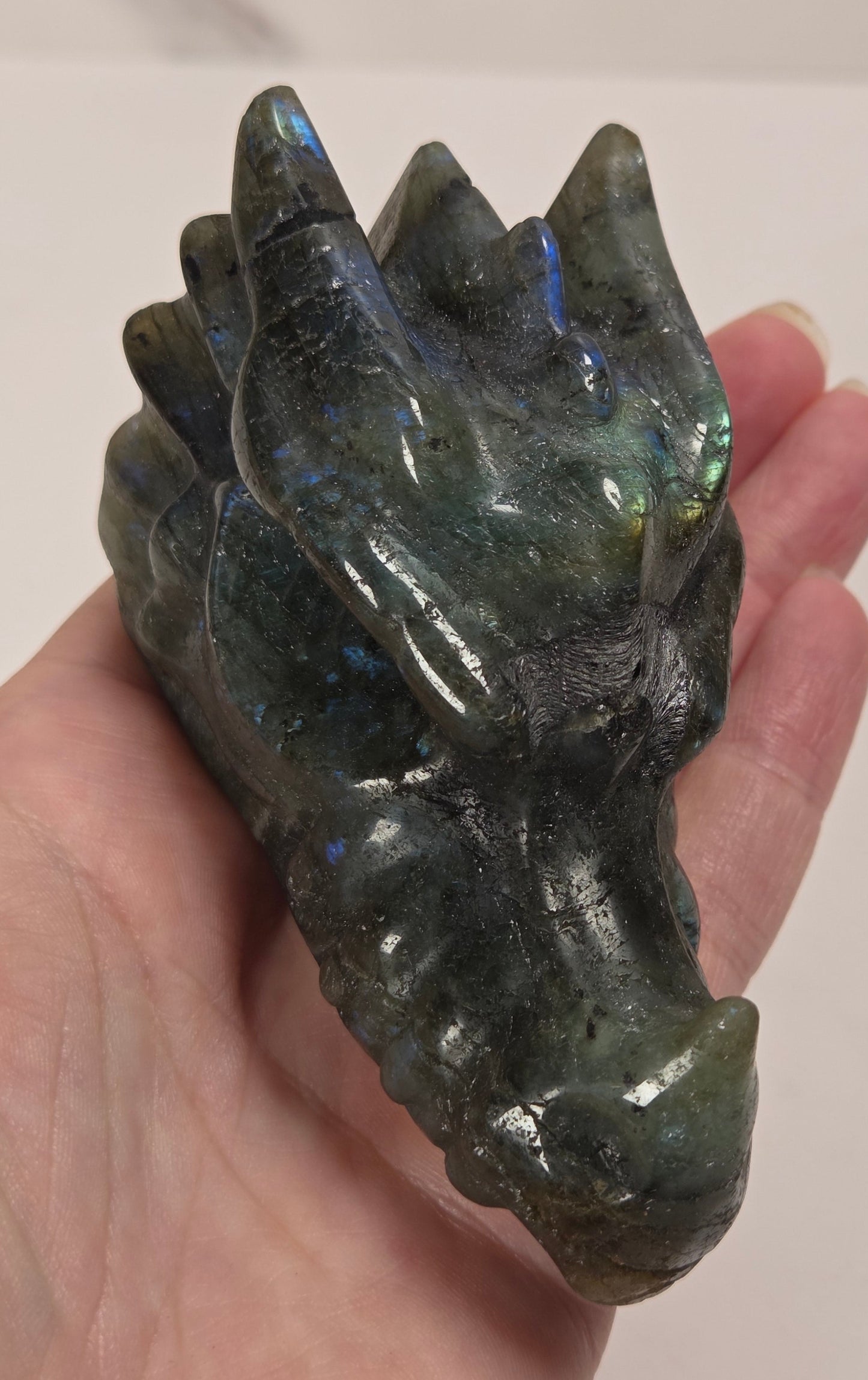 A hand holding a polished dark green labradorite crystal dragon head, showing its natural iridescent blue and green flashes, against a light off-white background.