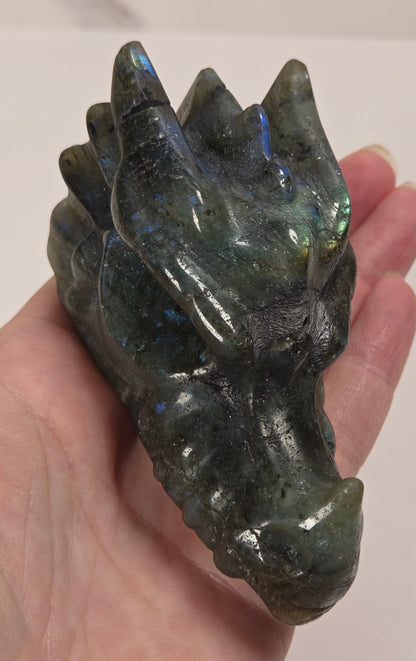 A hand holding a polished dark green labradorite crystal dragon head, showing its natural iridescent blue and green flashes, against a light off-white background.