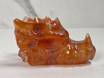 A carved dragon head figurine made of translucent orange-brown agate or stone, displayed on a light gray surface against a light gray background.