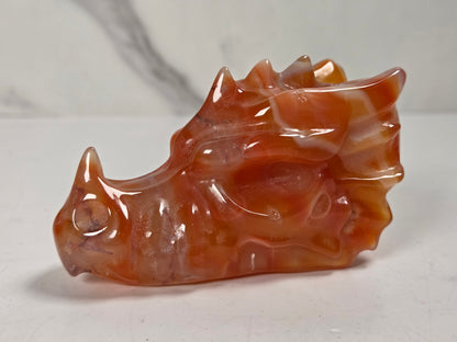 A carved dragon head figurine made of polished orange-brown agate, resting on a light gray surface with a marble-patterned background.