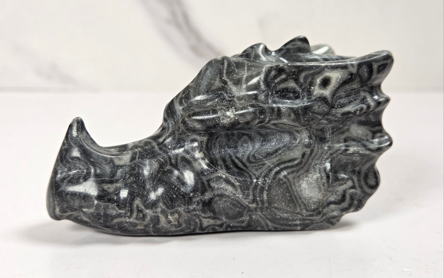 A carved black and gray stone dragon head figurine, showcasing natural marbled patterns, set against a light gray marble-patterned background.