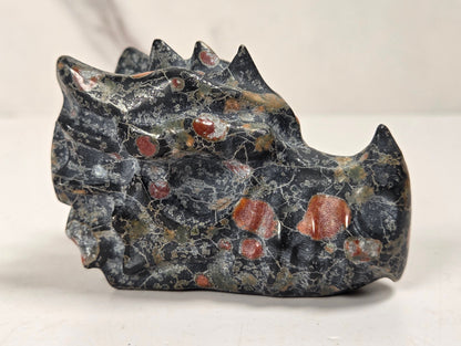 A carved stone object with a dark gray and black marbled texture and scattered reddish-orange inclusions, photographed on a light neutral surface.