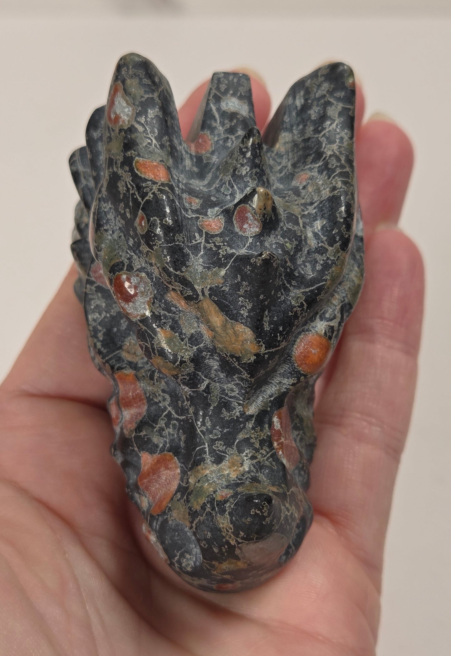 A hand holding a polished carved stone dragon head figurine. The stone is primarily dark gray and black with natural orange and brown inclusions.