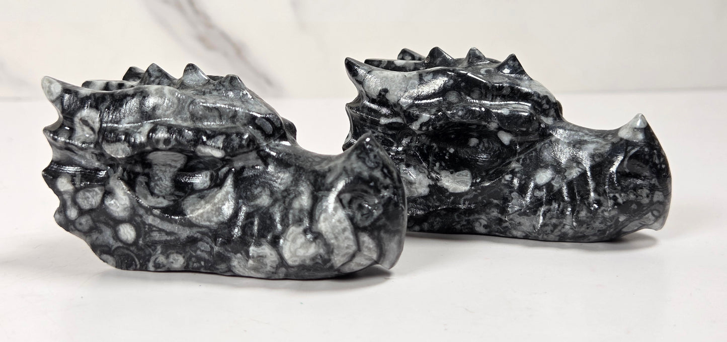 Two small carved stone dragon heads, likely made of marbled black and white stone, displayed on a white surface against a light gray marble-patterned background.