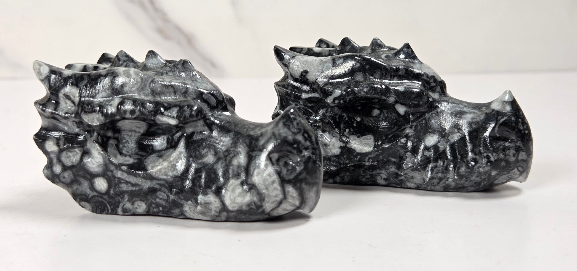 Two small carved stone dragon heads, likely made of marbled black and white stone, displayed on a white surface against a light gray marble-patterned background.