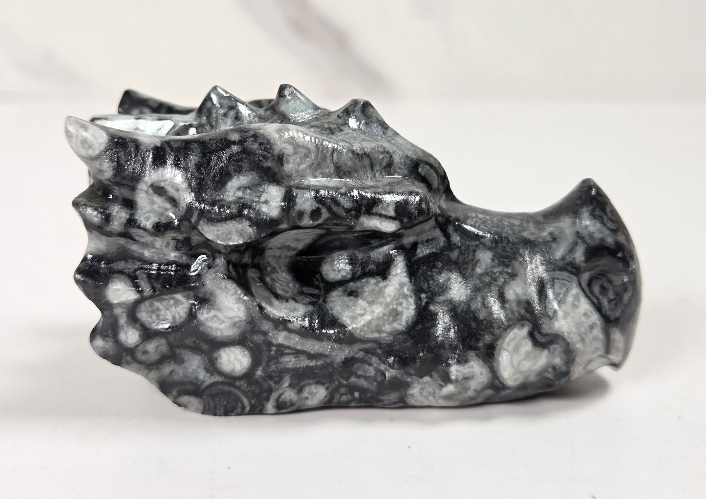A small carved dragon head figurine made of black and white mottled stone, shown on a light gray surface with a white marble-look background.