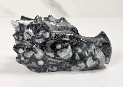 A small carved dragon head figurine made of black and white mottled stone, shown on a light gray surface with a white marble-look background.