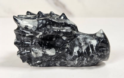 A carved black and white marbled stone dragon head figurine, displayed on a light gray surface against a white marble background.