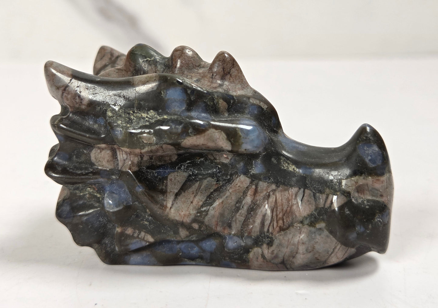 A polished stone dragon head figurine, showcasing natural inclusions of blue, brown, and gray, set against a light gray marble-patterned background.