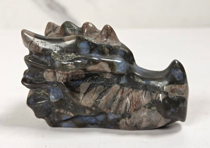 A polished stone dragon head figurine, showcasing natural inclusions of blue, brown, and gray, set against a light gray marble-patterned background.