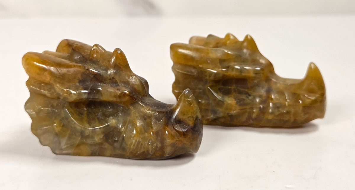 Two small carved animal head figurines, likely tiger or cat heads, made from polished brown and amber stone, displayed on a white surface with a light marble background.