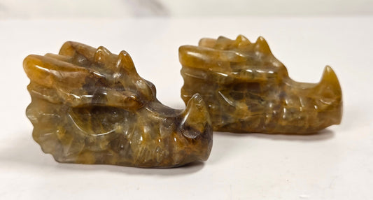 Two small carved animal head figurines, likely tiger or cat heads, made from polished brown and amber stone, displayed on a white surface with a light marble background.