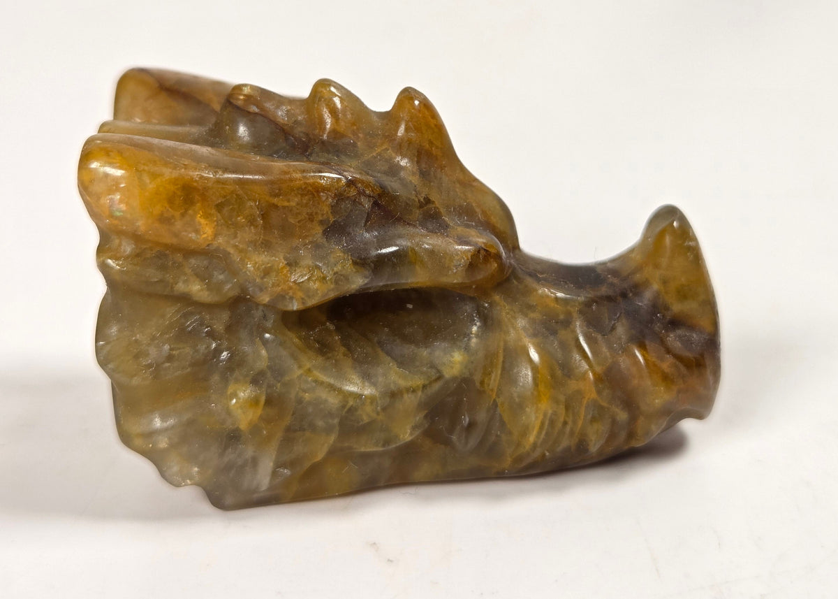 A small carved stone figurine resembling a dragon or animal head, made from polished brown and amber stone, photographed on a light neutral surface.