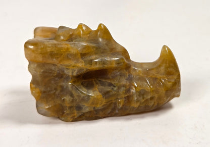 A small carved stone object with a rough, organic shape in warm golden-brown and amber tones, photographed on a light surface with a softly blurred light gray background.