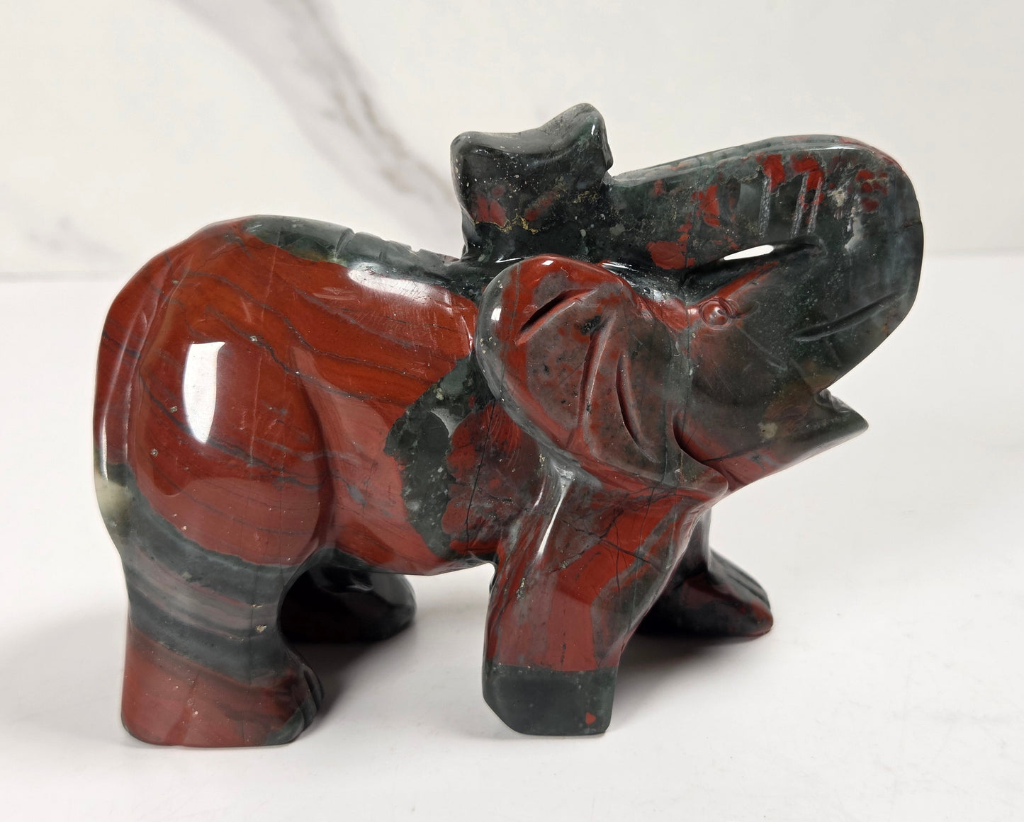 A small carved elephant figurine made from polished red jasper stone with dark gray to black veining, standing on a white surface against a light gray background.