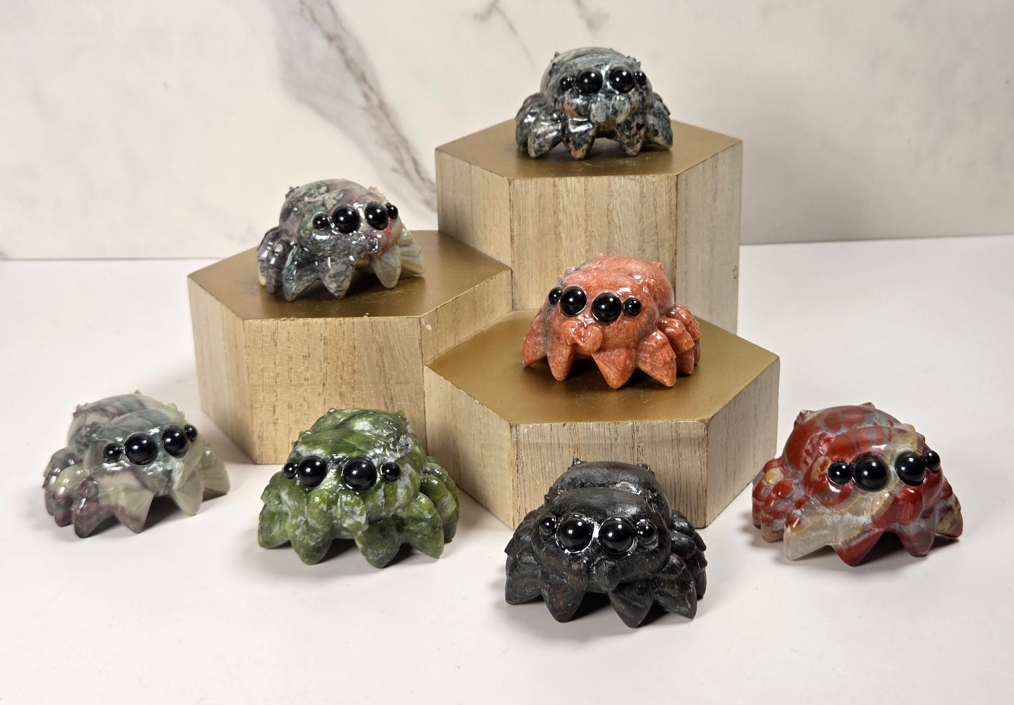 Seven small spider figurines in various earthy stone-like colors, each with large glossy black eyes, arranged on and around light wood hexagonal pedestals against a white marble-look background.