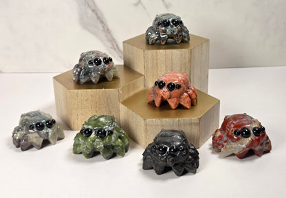 Seven small spider figurines in various earthy stone-like colors, each with large glossy black eyes, arranged on and around light wood hexagonal pedestals against a white marble-look background.