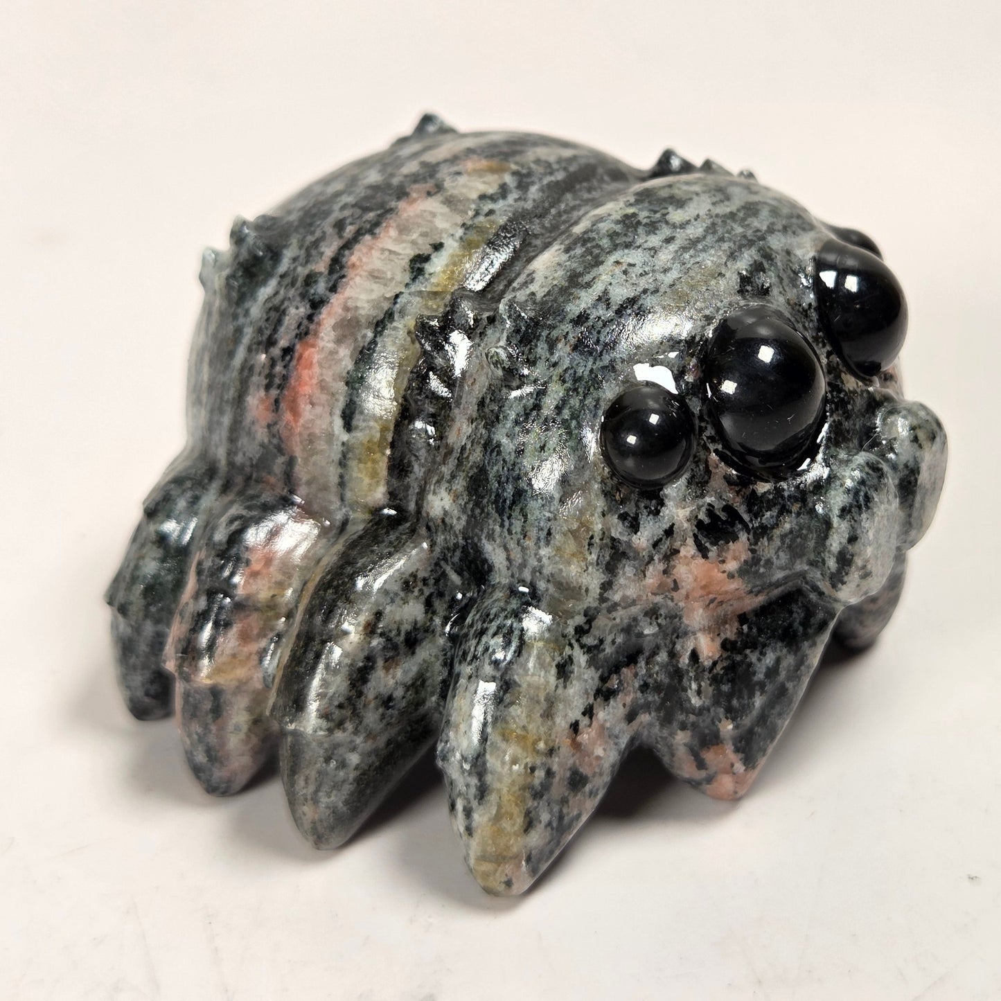 A small carved stone creature figurine with large glossy black bead eyes, featuring a mottled gray, black, and peach-colored stone body, set against a plain off-white background.