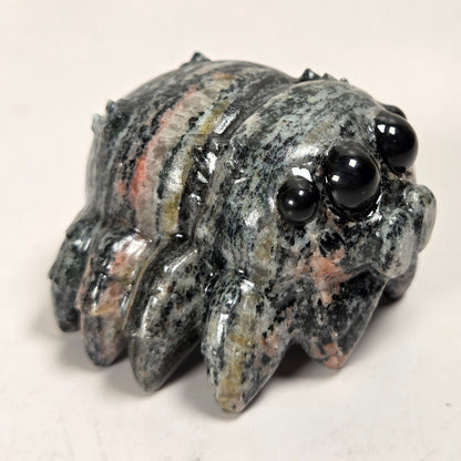 A small carved stone creature figurine with large glossy black bead eyes, featuring a mottled gray, black, and peach-colored stone body, set against a plain off-white background.