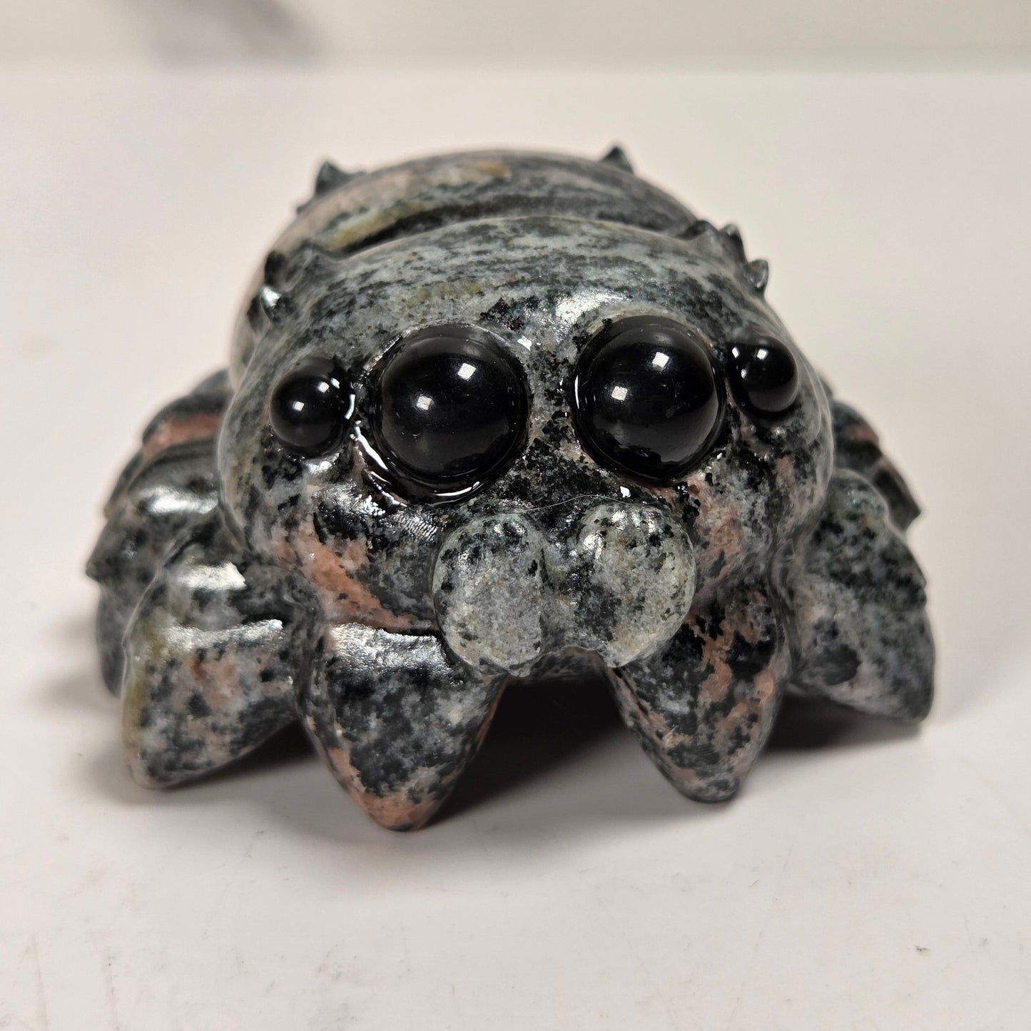 A small carved spider figurine made of speckled gray and black stone with glossy black bead eyes, photographed on a plain light surface.