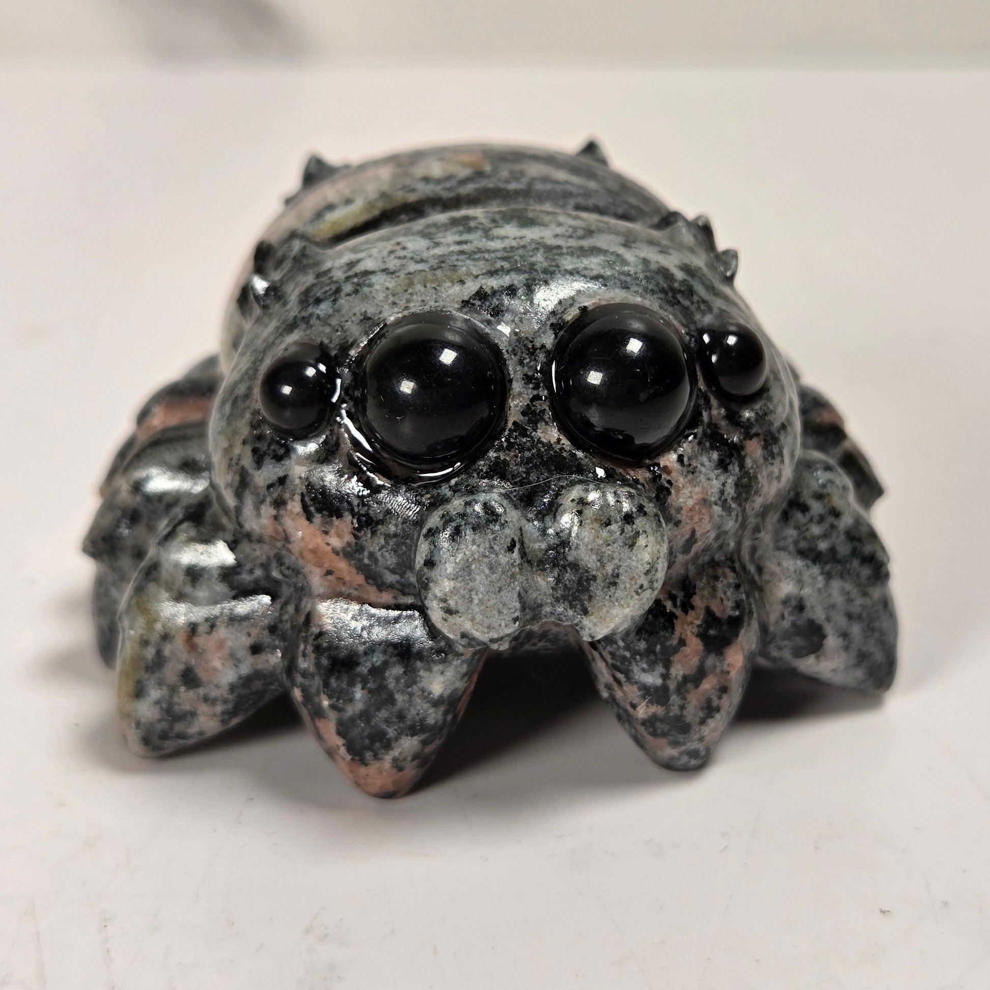 A small carved spider figurine made of speckled gray and black stone with glossy black bead eyes, photographed on a plain light surface.