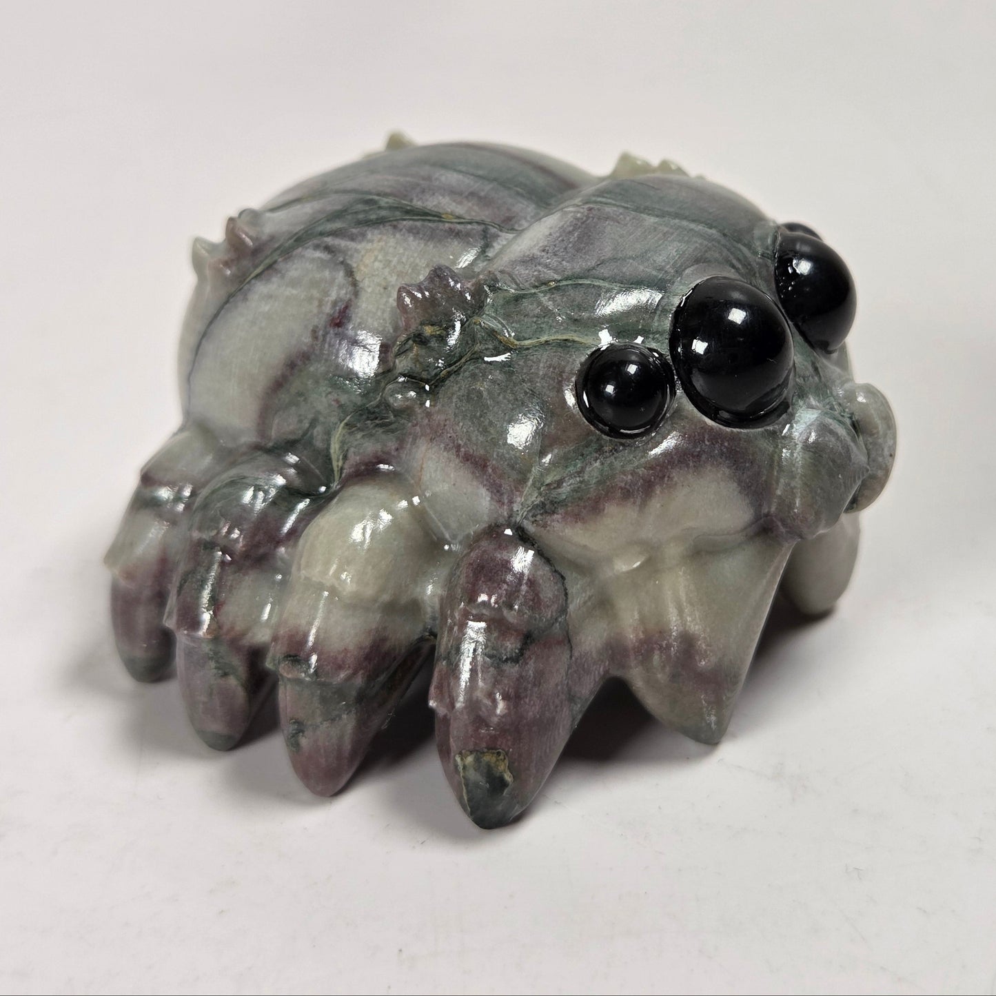 A small carved spider figurine made of polished gray stone with dark purple marbling and shiny black bead eyes, photographed on a plain light background.