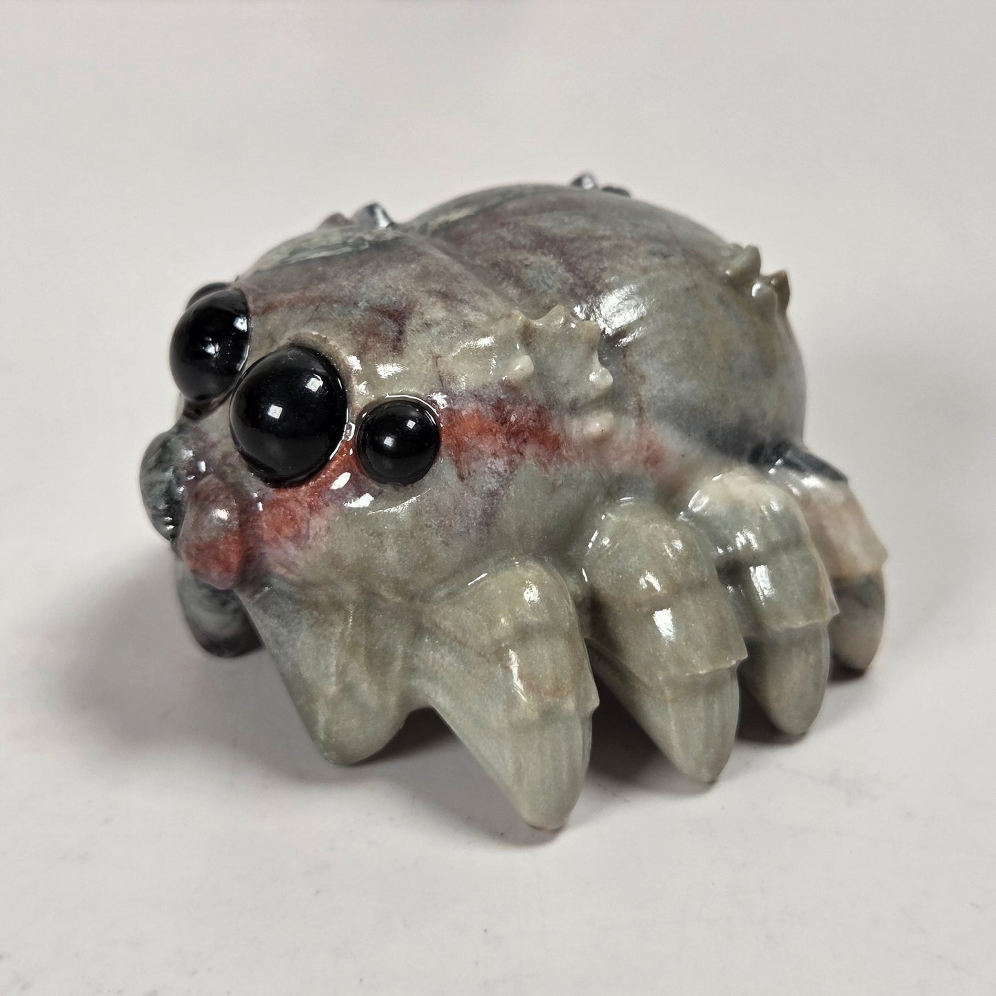 A small spider figurine with glossy black eyes and a gray body with subtle red markings, photographed on a plain light background.